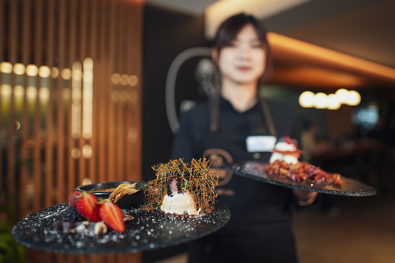 Eine freundliche Servicemitarbeiterin präsentiert im stilvollem Ambiente des Genussrestaurants mamazita zwei hochwertig angerichtete Desserts.